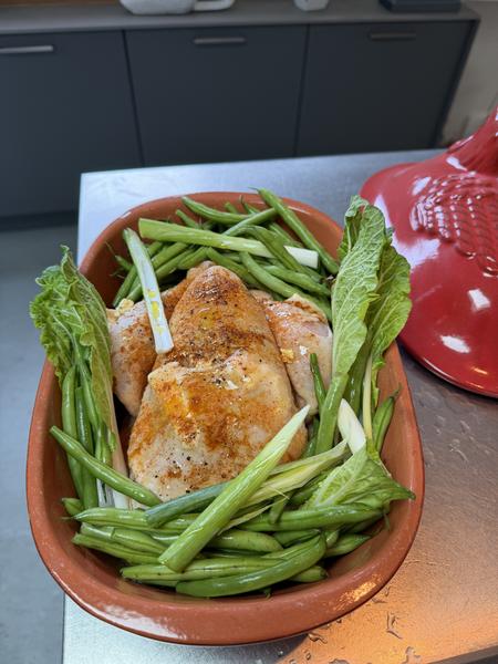 A whole chicken, seasoned and surrounded by fresh green beans and lettuce, rests in a terracotta dish on a kitchen counter